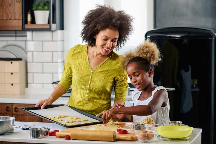 Smiling mother and daughter bake cookies together in a modern kitchen, using cookie cutters and a baking tray filled with dough shapes.