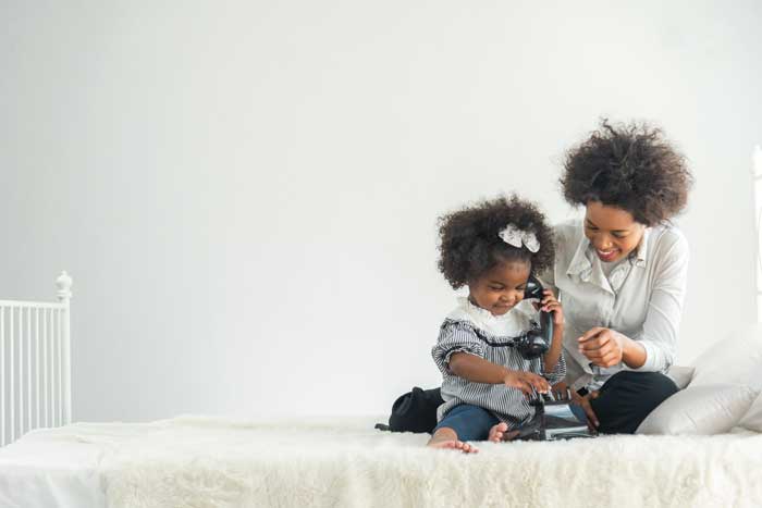 Smiling mother and her young daughter sit on a bed playing with a vintage telephone, enjoying a playful and warm bonding moment.