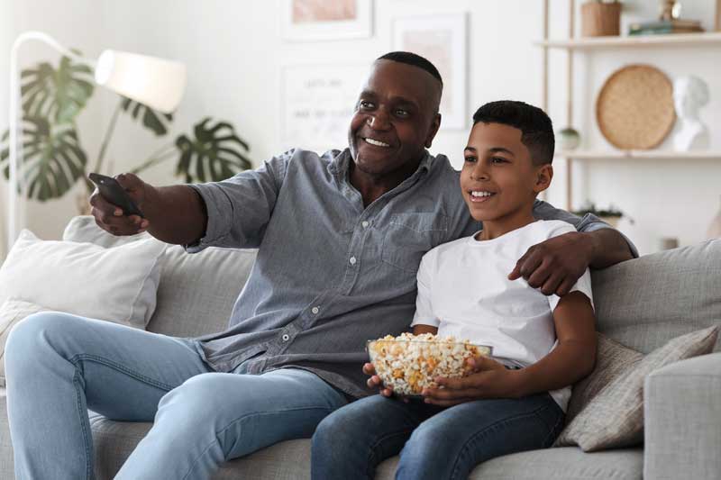 A smiling father and son sit on a couch watching TV, with the father holding a remote and the son holding a bowl of popcorn.