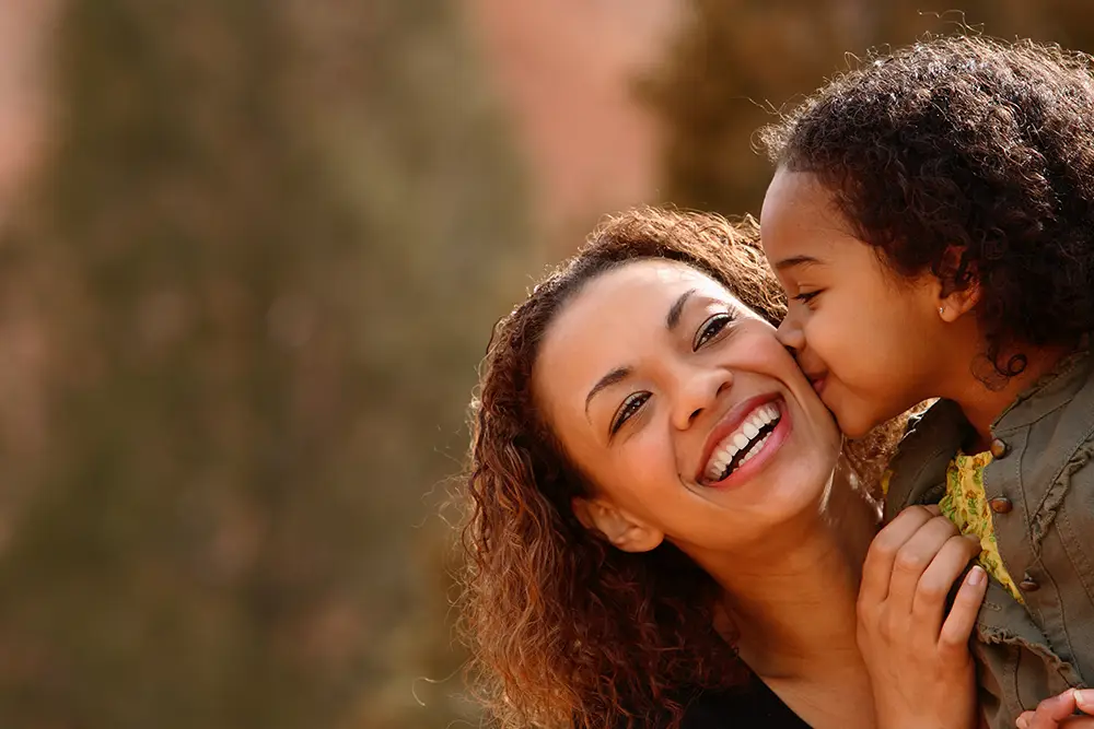A smiling mother being kissed on the cheek by her young daughter, symbolizing the deep bond between parents and children, even in complex custody situations.