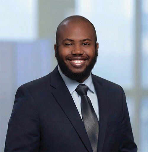 Professional portrait of a smiling Black man with a beard wearing a dark navy suit, light blue shirt, and black tie, against a blurred blue and gray background.