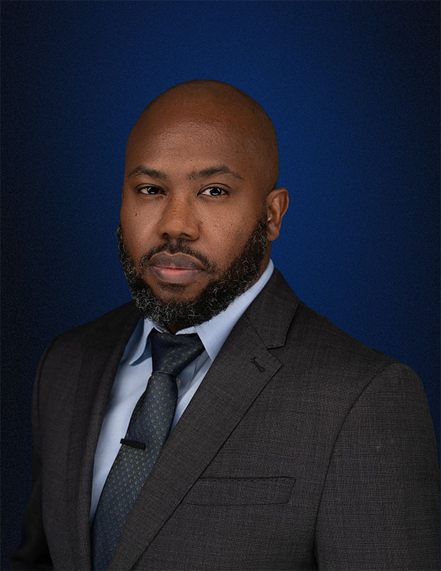 Professional headshot of Evander Williams, Esq., wearing a charcoal suit and tie against a blue background.